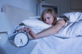 Displeased young woman in bed extending hand to alarm clock at home